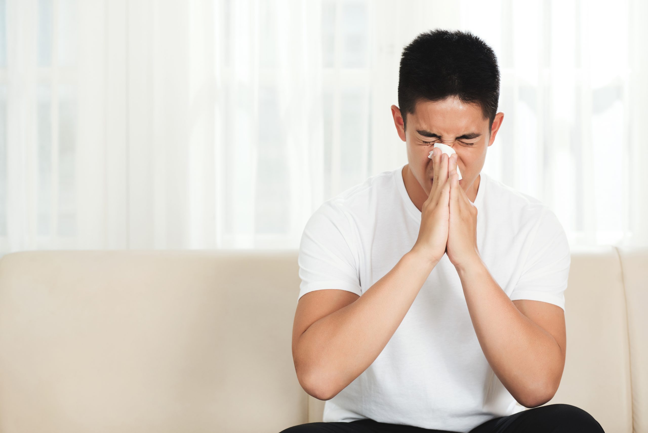 young-asian-man-sitting-couch-home-blowing-nose-with-tissue-scaled